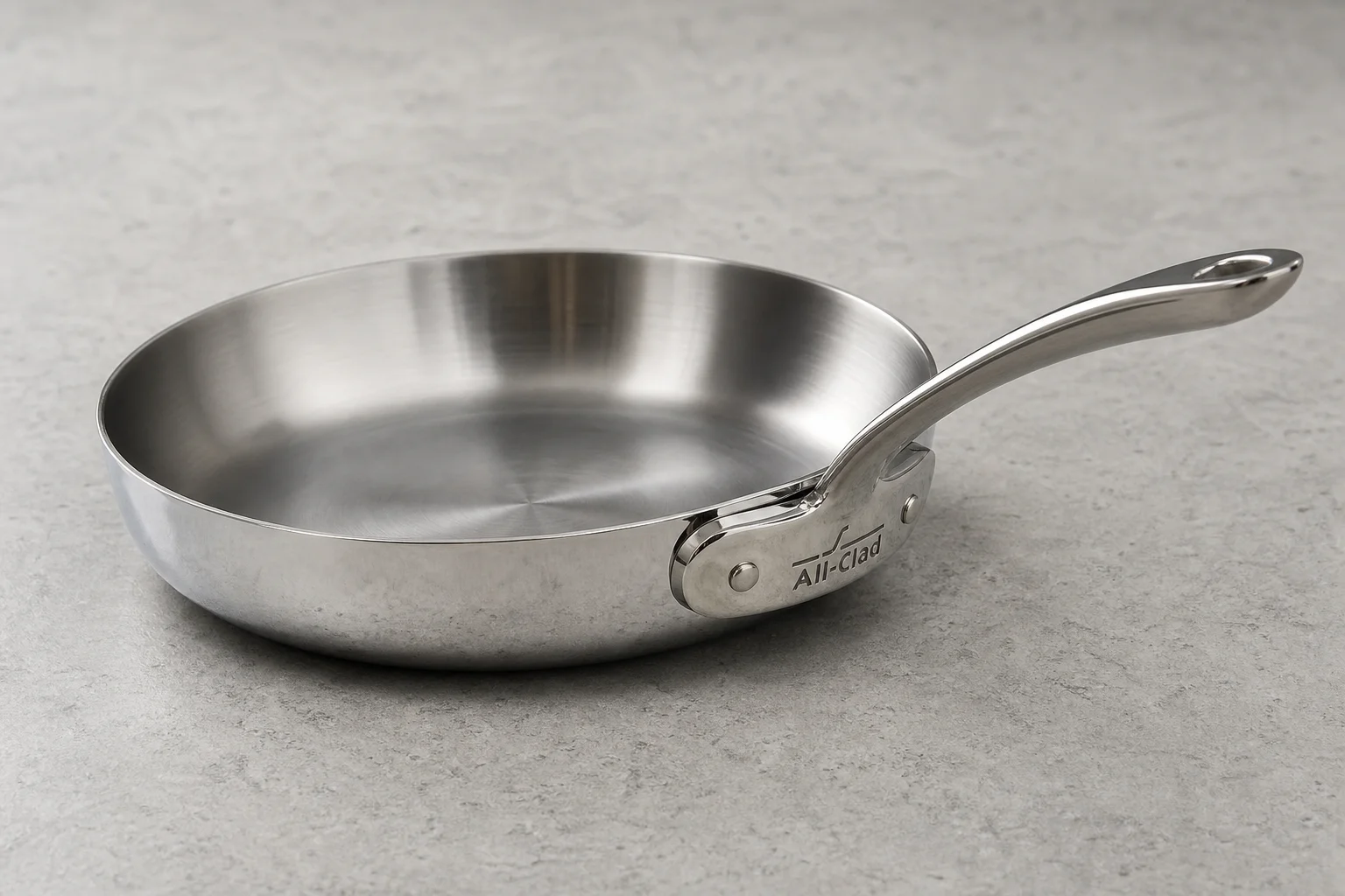 Collection of six stainless steel frying pans with varied finishes and handle styles on concrete surface
