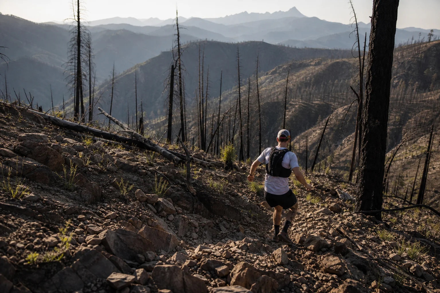 Trail runner descending steep rocky burn area with charred trees and loose terrain