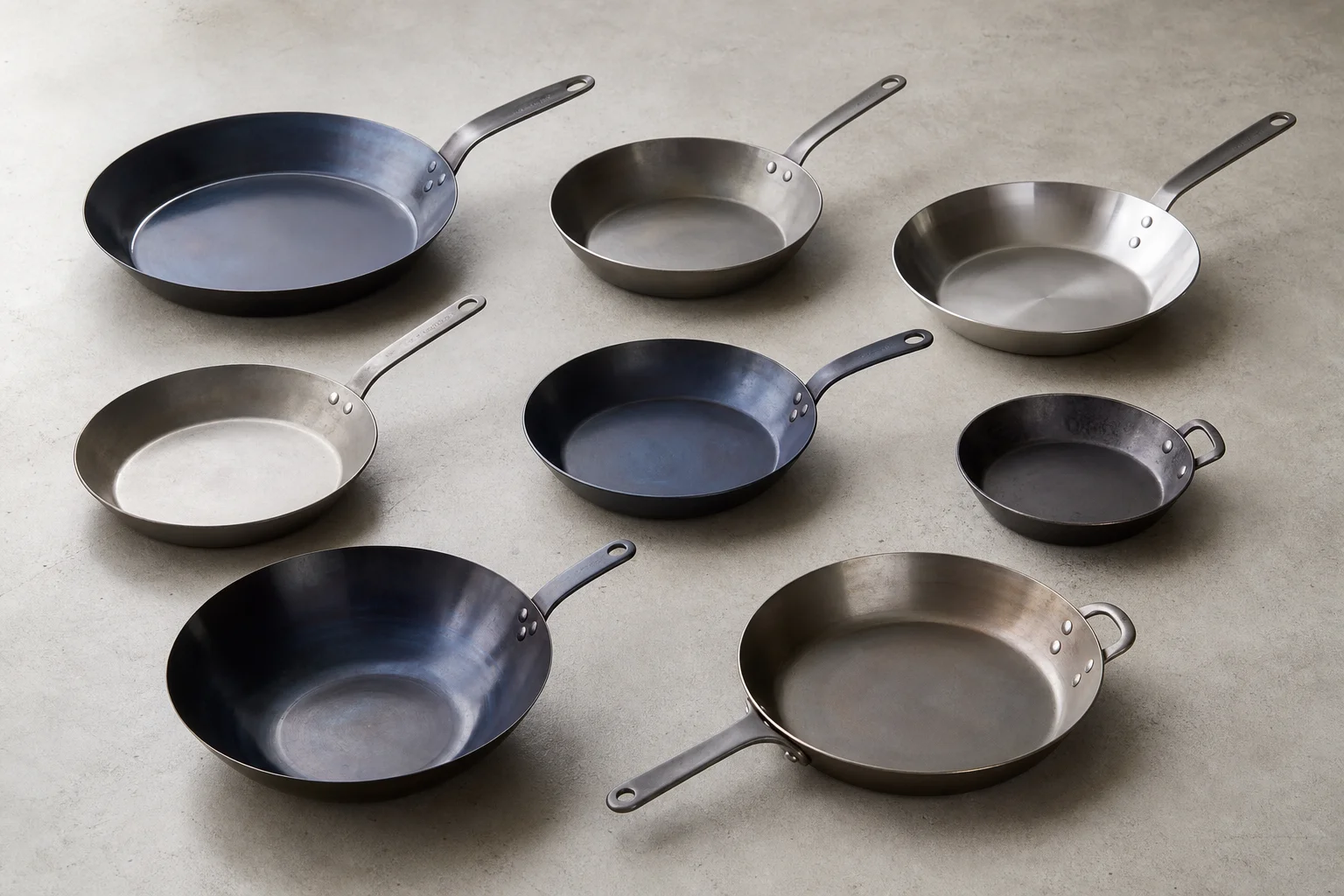 Eight carbon steel pans in various colors and sizes arranged on concrete
