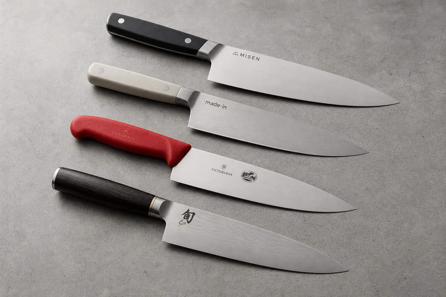 Four chef's knives arranged diagonally on concrete: Misen, Made-In, Victorinox, and Shun