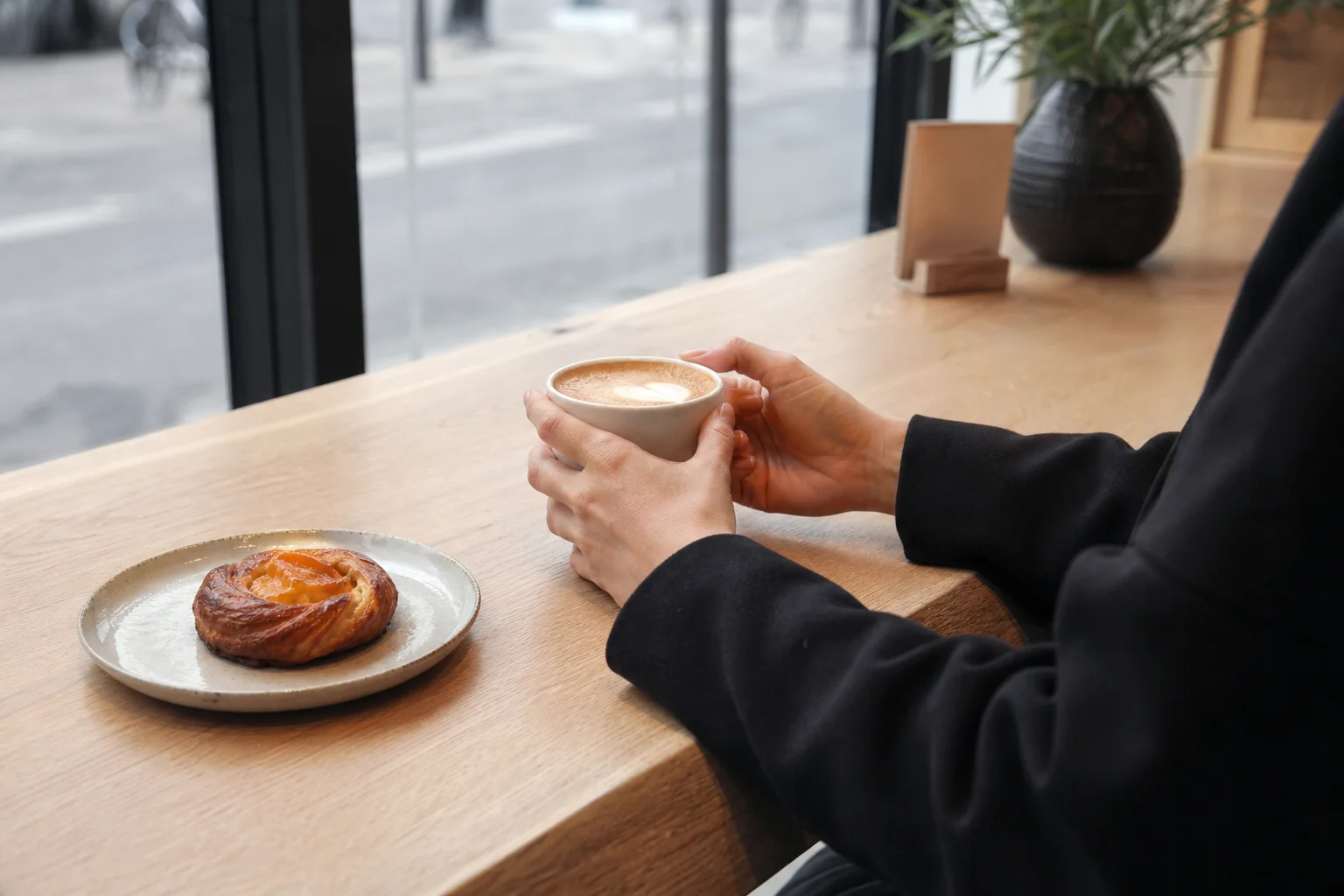 Person at specialty cafe counter with cappuccino and pastry, city view through windows