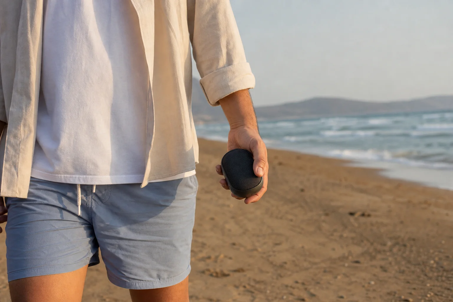Person holding compact speaker at beach with ocean waves