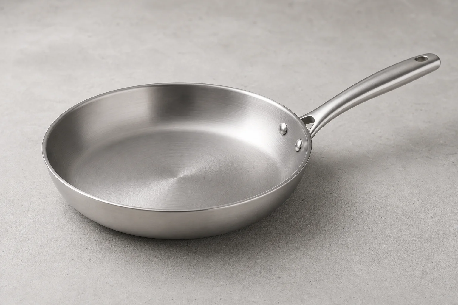 Collection of six stainless steel frying pans with varied finishes and handle styles on concrete surface