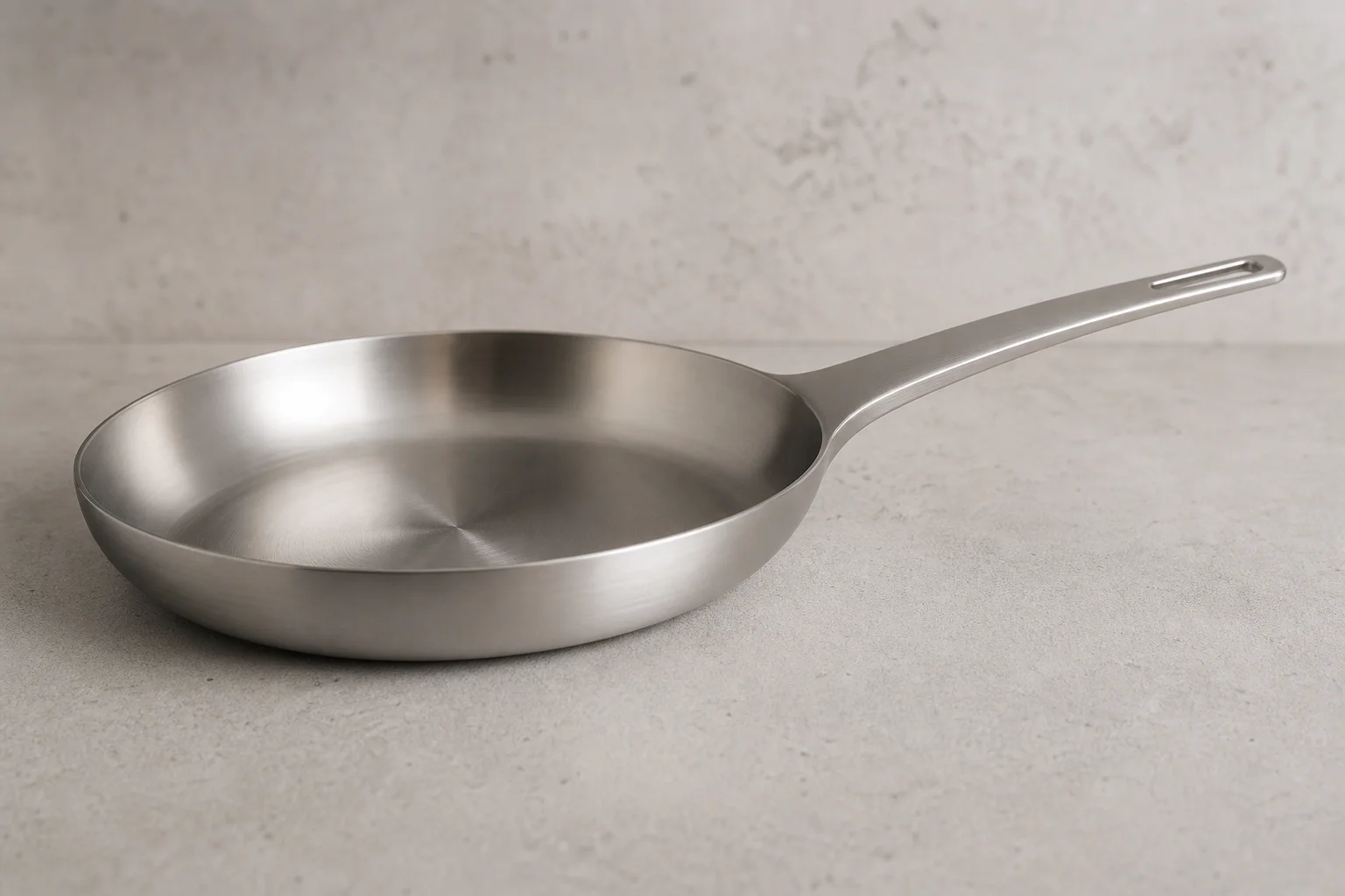 Collection of six stainless steel frying pans with varied finishes and handle styles on concrete surface