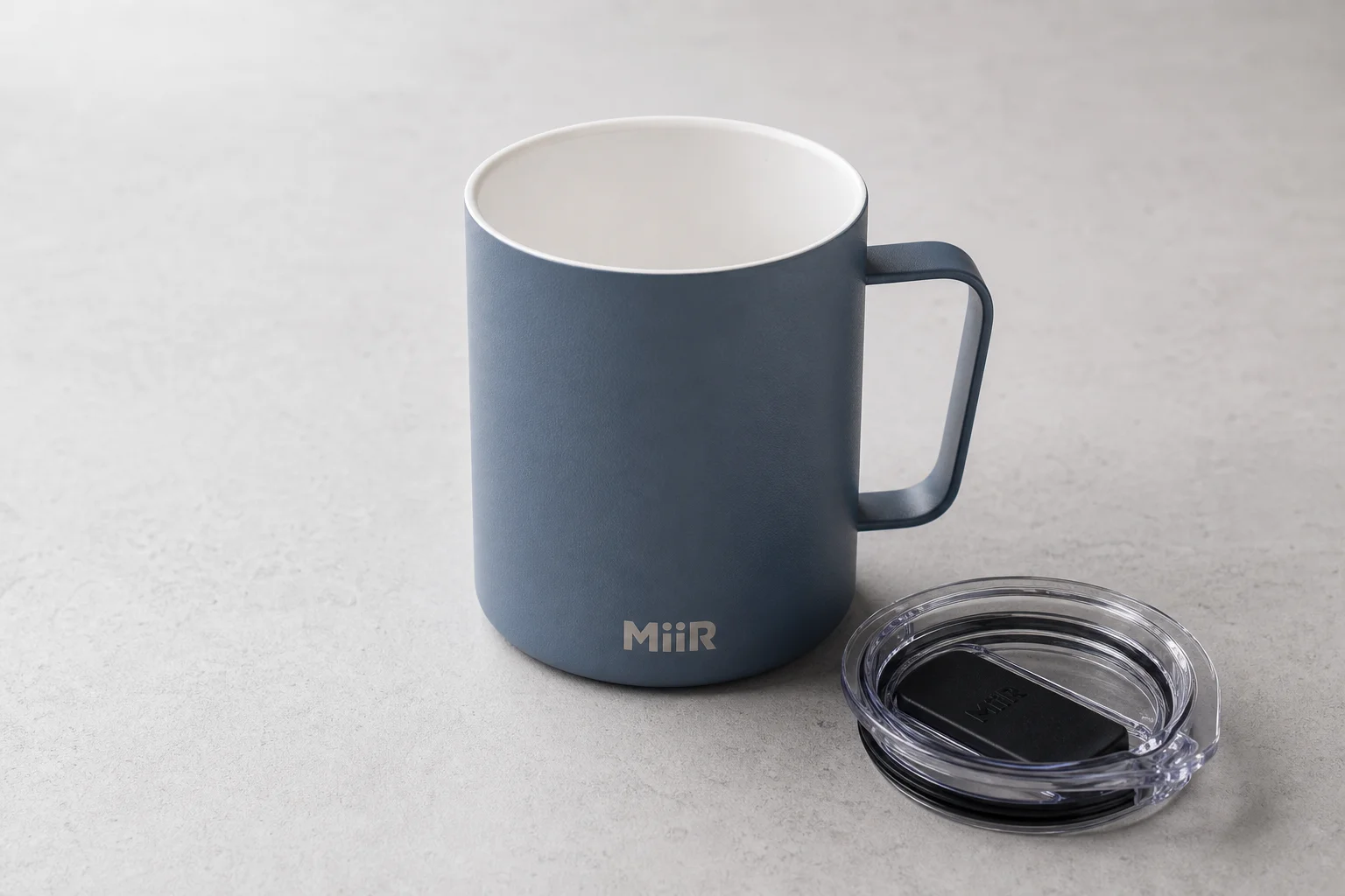Eight reusable coffee cups arranged on concrete surface showing variety of materials and designs