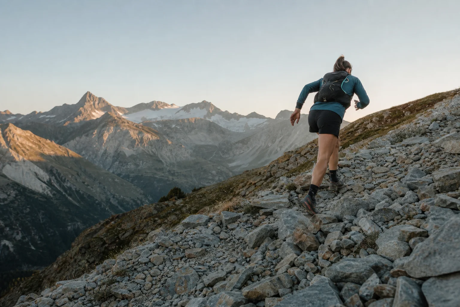 Trail runner ascending a rocky alpine slope with distant peaks in soft morning light
